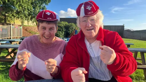 BBC News Gloria and Jayne sit at a picnic table clasping their fists in a show of celebration and hope, wearing their caps