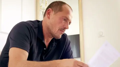 A man with short, light brown hair and stubble looks at a piece of paper. He is wearing a dark blue, short-sleeved polo shirt.