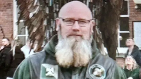 James Martin, who has a long grey beard and is wearing a leather biker gilet and green jumper, looks into the camera