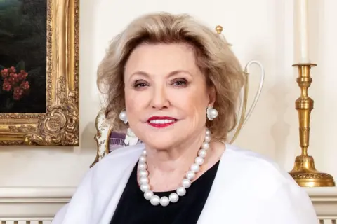 Reuters Barbara Taylor Bradford is photographed against what looks like an opulent fireplace. She has blonde hair and her smile reveals very white teeth. She is wearing a cream jacket over a black top and has matching pearl earrings and necklace.