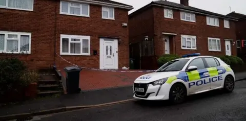 A police car is parked on the road outside a house, where police crime tape can be seen cordoning off the brick-paved driveway. The house has a white door and a white-framed front lower window.