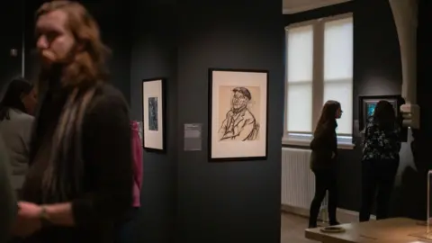 The Auckland Project The drawing framed and on display in the exhibition gallery. People are milling around looking at paintings.