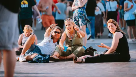 Nadine Ballantyne Two women and a man sat down at Bristol Sounds. They are all looking at the camera. The man and woman are pointing at the camera while the other is holding her phone and looking at the lens.