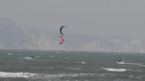 Mandy Lennox-Gordon Two kitesurfers in a rough sea, with the coastal cliffs in the background. One kite is mainly red, the other blue.