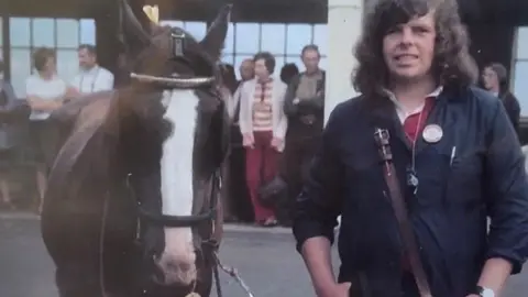 Isle of Man Transport An old photo Peter Cannon, who has long brown waving hair, standing next to a brown and white horse.