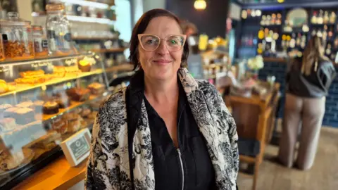 Stuart Woodward/BBC Tracie Sullivan stands in front of a counter in her cafe containing cakes and other food items. She is wearing a white jacket with navy floral detail, over a black top. She has peach-framed glasses on and is smiling at the camera. 
