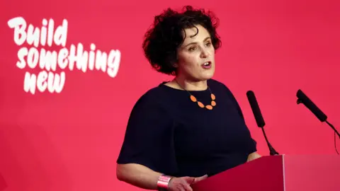 Claire Hanna, wearing a navy top, standing behind a red podium with two microphones on it. The wall behind her is read with "build something new" written in white. 