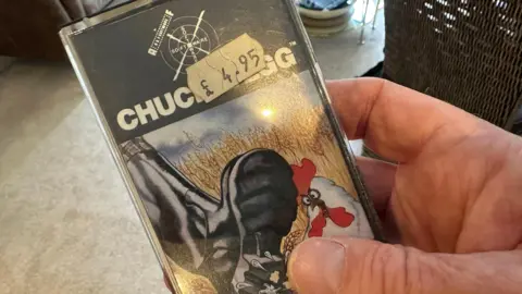 A hand can be seen holding a card with the words "chuckie egg" and a £4.95 price sticker stuck on it. There is a cartoon image of a black boot about to stamp on a chicken on the card.