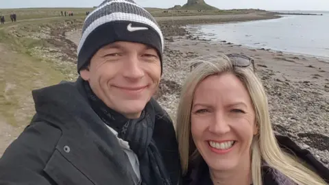 A man wearing a Nike beanie hat and a big coat and a woman with long blonde hair and glasses on the top of her head smile at the camera. They are standing on a beach.