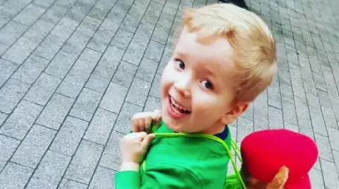 A boy with blonde hair, a green top and a red bag on his back standing on grey paving