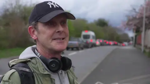 The image shows a man wearing a dark New York Yankies baseball cap and some headphones round his neck. He stands beside a road with a tailback of traffic in the distance.