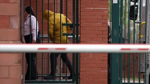 A man in a yellow hoodie pulled over his head and black jeans walking from a custody van inside a barred doors on a red brick building. A woman custody or court officer in a white top and black bottoms is holding the door