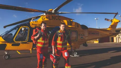 Lincs & Notts Air Ambulance Two men dressed in red overalls with yellow fluorescent stripes on them are carrying paramedic items and walking away from a yellow Lincs & Notts Air Ambulance helicopter that is parked on tarmac.
