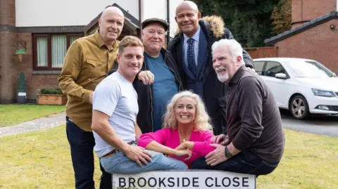 Lime Pictures Paul Usher, John McArdle, Michael Starke, Suzanne Collins e Philip Olivier são retratados com a placa de rua Brookside Close. Eles estão todos sorrindo para a câmera. Uma casa com gramado na frente e um carro branco estacionado na garagem são visíveis ao fundo