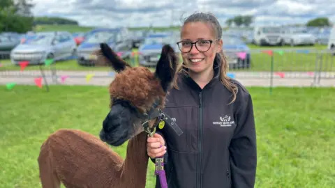 Beth Hobson, a woman wearing glasses and a navy blue jacket, holding the reins of a brown alpaca, is standing in a field. There are cars parked in the distance behind.