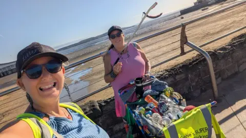 Two women photographed at the beach, with a trolley full of collected waste. One is holding a litter grabbing tool