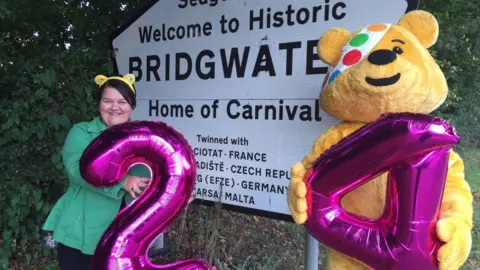 Emma pictured left, wearing yellow Pudsey ears and a green jacket. She is standing by a 'Welcome to Bridgwater' sign with a pink balloon in the shape of the number two. Pudsey stands on the right holding a pink balloon in the shape of the number four.