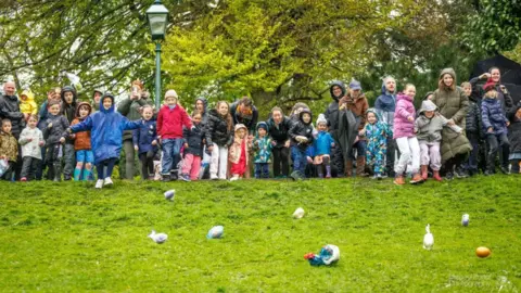 Micheal Porter Photography A group of people wearing coats and winter clothing stand at the top of a grassy hill, with large, designed eggs rolling down it. Some of the eggs are in plastic bags. There are leafy trees and a lamppost in the background.