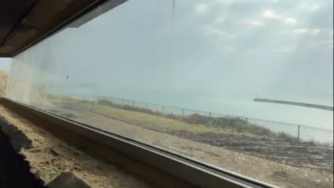 George Carden/BBC The battery observation post is a concrete bunker with a panoramic window which gives a view of the cliffs at Seaford, the ocean and Newhaven Harbour. Grass and mud are visible at the front of the viewing point which is almost level with the ground and near the edge of the cliff edge.