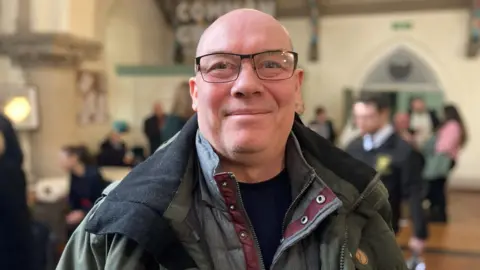 Mr Hunter is smiling for the camera, inside the church with the pews in the background.  He has a bald head and is wearing a green coat with glasses.