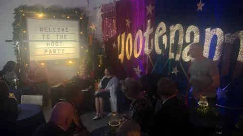 A group of people are gathered around tables and sat on a sofa. There is a sign on the wall which reads 'welcome to the hoot party' and there are glittering letters on a purple curtain which read 'Hootenanna'.