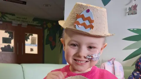 Amy Dixon A picture of Freya in a hospital setting, smiling with a sick bowl decorated with an easter egg, that she's wearing as a hat. She has a tube in her nose and is wearing a pink jumper.