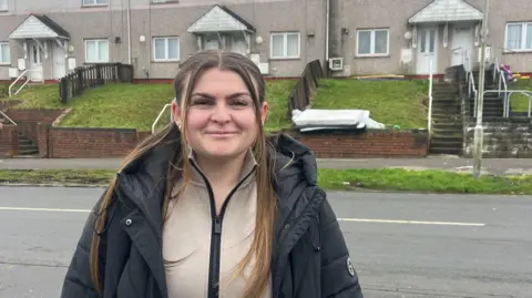 Taylor Richards who has long brown hair and is wearing a beige jumper and black coat, stood in front of Gors Avenue