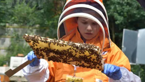 A young girl wearing an orange beesuit holding a frame of honeybees and a hive tool in her other hand. She is wearing blue gloves.