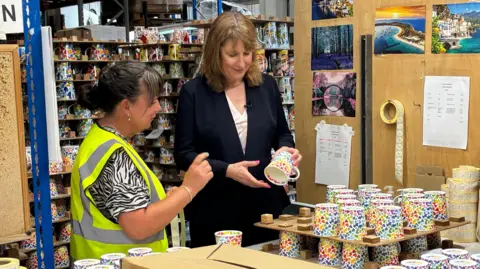 LDRS Rachel Reeves, wearing a dark suit, smiles as she holds and examines a mug while in a ceramics workshop. A woman wearing a hi-vis jacket and standing next to her is pointing at the mug explaining something to Reeves.