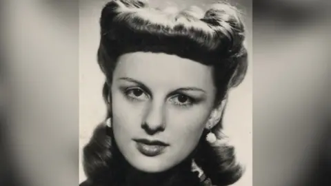 A black and white headshot image of Carol in her twenties looking down the lens of the camera. She is wearing drop white ball earrings and her hair is highly styled.