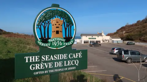 National Trust Jersey A large, bold, green 1936 Jersey national trust sign with a picture of a castle. The words 'The Seaside Cafe' can be seen with a moderately busy carpark in the background. The sky is blue with no clouds in the distance.