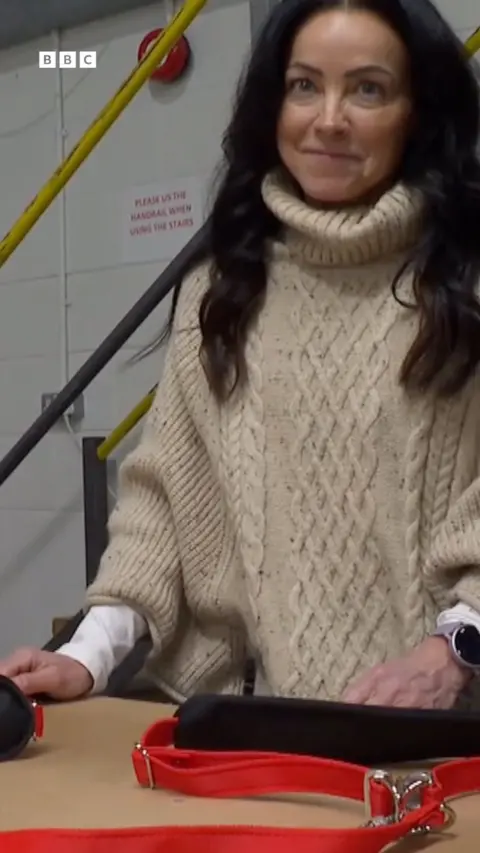 A woman with brown curly hair, wearing a cream knitted sweater smiles into the camera. Below her on presented on a table is a red and black harness.