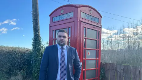 Alan Lewis Alan Lewis in front of a red phone box. He has dark hair and beard, wearing a dark blue blazer, light shirt and check tie.