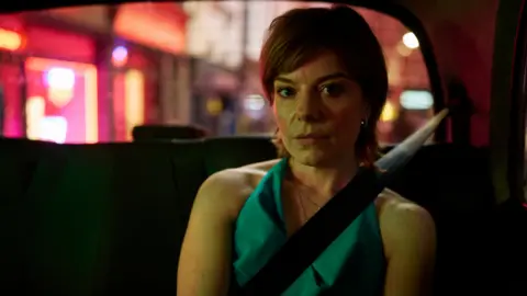 ITV The image shows a young woman sat in the back of a car with city lights illuminating the car. She has short ginger hair, she is wearing a green top and her seatbelt