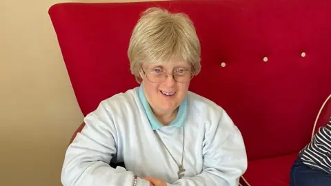 SUPPLIED Woman with short blonde hair in white jumper sat on red sofa