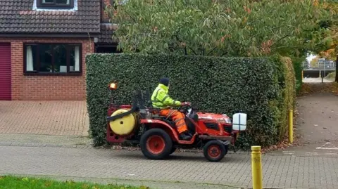 Judith Heinemann A red mini-tractor being driven by a man in yellow and orange PPE. There is a yellow barrel attached to the back of the vehicle. It is driving past a green hedge, with a block-paved drive to the left and the edge of a two-storey house.