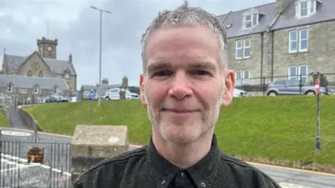 A man with short grey hair and stubble in a black shirt buttoned up at the collar stands in front of an expanse of grass with buildings behind