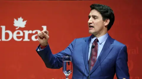 Getty Images Justin Trudeau stoi na podium przed czerwonym tyłem. Ma na sobie niebieski garnitur i wzorzysty krawat i wskazuje jedną uniesioną ręką.