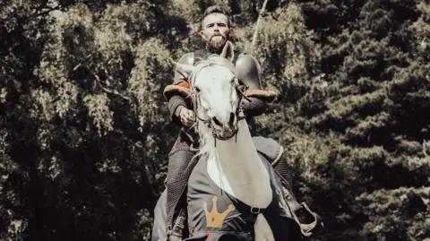 Henry Browne - a man dressed in a medieval knight uniform - has brown hair, a moustache and beard and is looking directly at the camera with a serious expression whilst he is sitting on a white horse. Trees can be seen behind him.