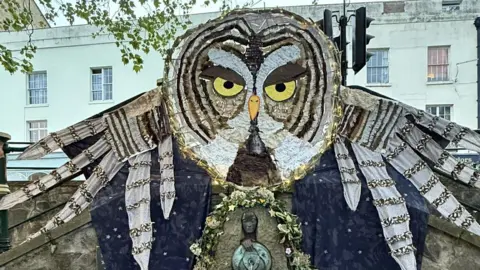 A huge tawny owl figure has been attached above a small spring well set in a stone wall. The owl has large yellow eye and its wings are spread as if in flight