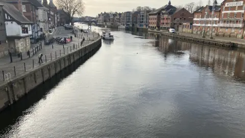 The River Ouse in York, pictured from a bridge. A boat can be seen in the distance. Properties flank the river on both sides.