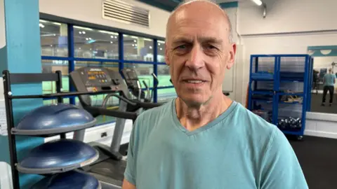Anthony is stood in one of the gyms at Cardiff Met university, where he continues his physiotherapy since his cardiac arrest. Behind him are running machines and free weights. He is wearing a green t-shirt and looking at the camera.