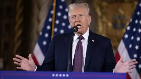 Donald Trump gestures with his hands during a speech. Behind him are US flags