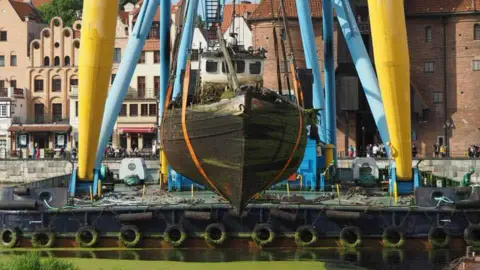 Maritime Heritage Trust A head shot of a rotten wooden fishing boat being lifted out of a marina. There are two yellow legs of the crane in the background and a lot of green algae in the water