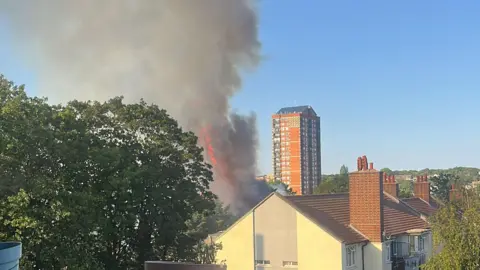 MICHELLE BERRIER A view from a balcony showing a large column of thick grey smoke and orange flames rising from a fire behind trees in a residential area. A tall brick tower block stands in the background under a clear blue sky, with a quiet street, a bus stop, and a parked car visible below.
