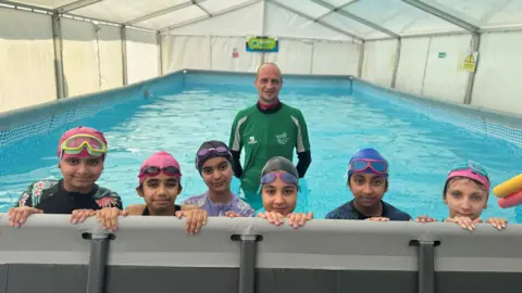Six children are in a swimming pool with swim caps and goggles on their heads. Their is a swim instructor behind them. They are in a marquee. 