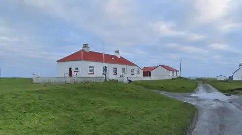 A white building with a red door and red roof. The building is surrounded by grass with a single track road running down the right hand side. The sky is dappled with clouds.