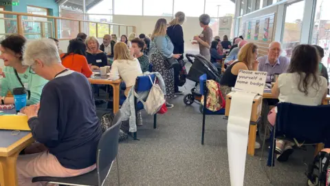 Educafe A room in a library with lots of people sitting round small tables chatting