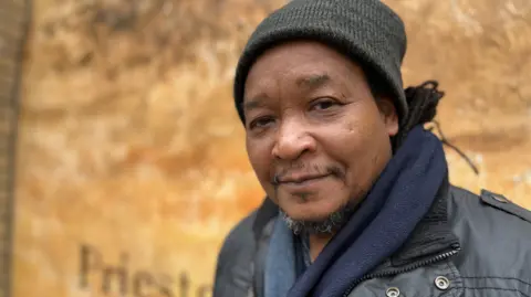Emma Baugh/BBC Tony, a man, wearing a grey woollen hat, with a small grey beard. He is wearing a blue scarf and a grey jacket, smiling for the camera.
