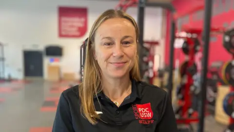 Dr Kate Williams has long brown hair. She is smiling at the camera, wearing a black polo shirt. There is red logo of University of South Wales staff on the right shoulder of her top.